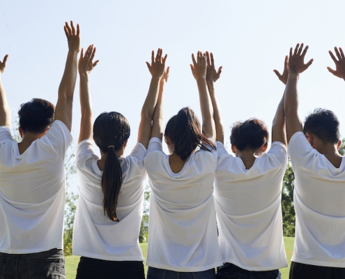 Happy excited young people in white t-shirts raising arms when standing in city park, view from the back